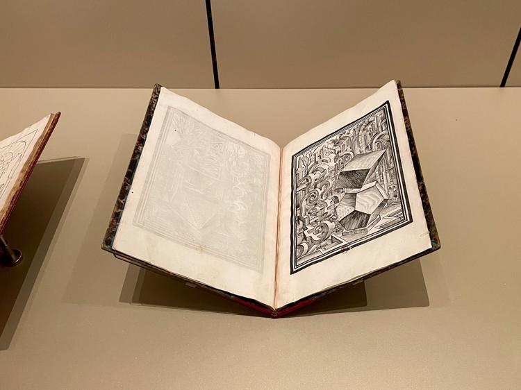 A close up of a book in a display case. The left-hand page is blank, while the right-hand page has a black-and-white line drawing of fantastical geometrical shapes, vaguely reminiscent of an Escher drawing.