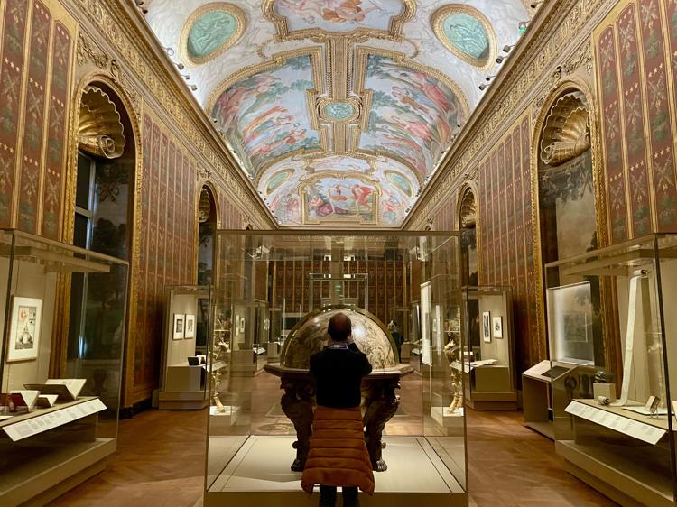 A long, ornately decorated gallery with gold trim on the walls and colourful paintings on the ceiling. There are various display cases against the walls, and directly in front of me is the silhouette of a man standing in front of a large globe.