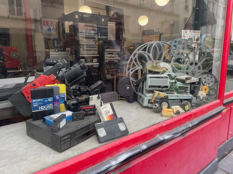 A window display with old video gear tastefully arranged -- including a VHS player, various tape cassettes, a large reel-to-reel film projector, and a few video cameras.