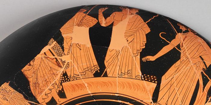 A black and orange fragment of vase depicting a scene of two men voting at an altar. A woman stands on the altar, with a pile of small circles to represent pebbles on either side. On the right-hand side, a man is reaching down to put a pebble on one of the piles and cast his vote.