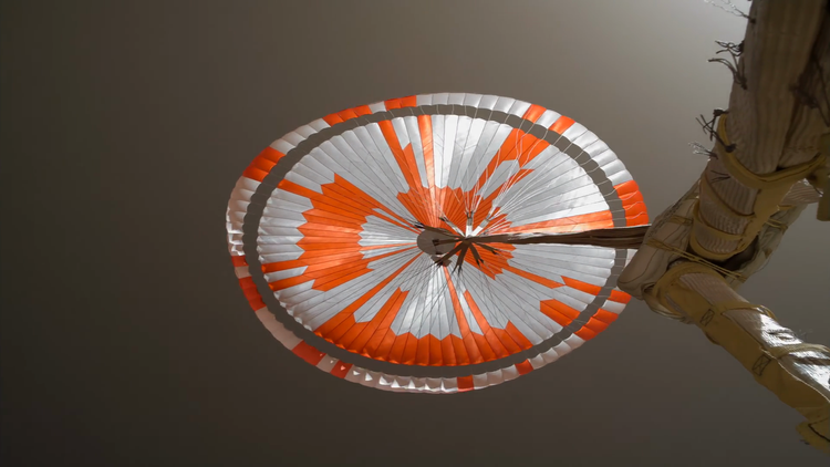 Photo looking up towards a parachute against a dark brown sky. The parachute is made of white-and-orange segments, and is stretched horizontally. The circle is wider than it is tall.