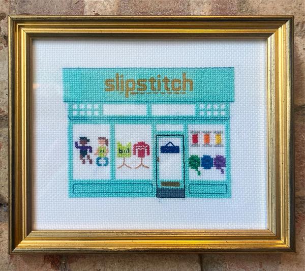 A rendition of the shopfront in cross-stitch, mounted in a gold frame on a brick wall. The shop is a simple geometric design in turquoise thread, and each of the three windows shows a mini-display – a pair of barbies, a pair of jumpers on stands, six balls of wool in rainbow colours.