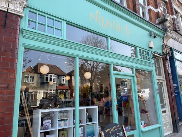 A photo of the shop's exterior. It has bright turquoise paint with the word ‘Slipstitch’ in gold lettering across the top, and large windows that show the shop interior. You can see shelves, balls of wool, and two large wooden knitting needles.