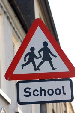 A warning sign labelled ‘School’. The sign is a red triangle with a white background, and two stick figure children are walking from left to right. The right-hand child is slightly older and walking the younger child by the hand. Their heads are attached to their bodies, and the older girl’s head is non-circular, but in a subtle way.