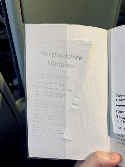 A faded piece of paper tucked inside a book, titled 'Hertfordshire Libraries' and with details of the loan, including the loan period and library contact details.