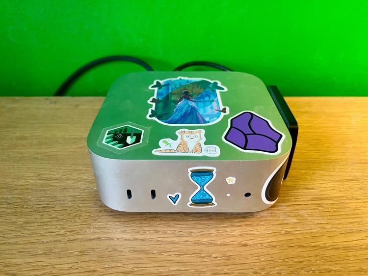 A Mac mini covered in stickers with a distinctly green tinge, reflected off the green wall behind it.