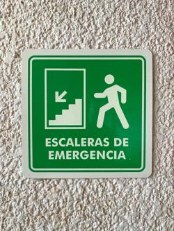 A green sign with white lettering that says ‘Escaleras de emergencia’ with a stick figure of a running person. The stick figure’s head is detached from their body and a perfect circle.