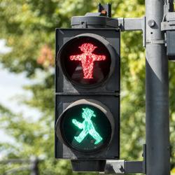 Red and green pedestrian crossing lights. The red light is a small figure with their arms oustretched and a wide brimmed hat, while the green light is the same figure walking, their head tipped back slightly.