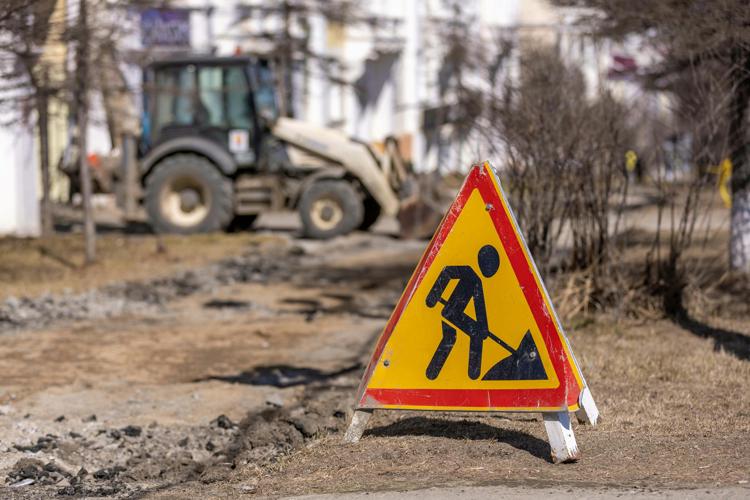 A construction site with a warning sign in the foreground. The sign is a red triangle with a yellow background, and a black stick figure shovelling something. The stick figure’s head is separated from their body, and a sort of oval shape.