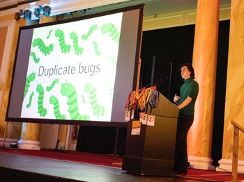 A person in a green top standing at a podium, in front of a large screen with the words “duplicate bugs”.